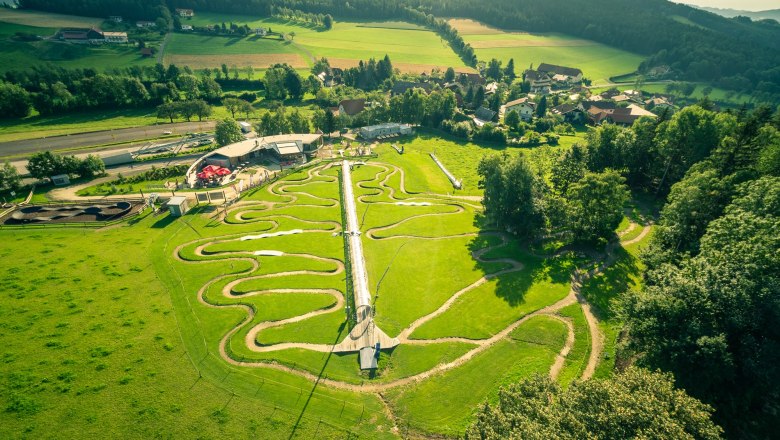 Die Wexl Arena St.Corona, © Wexl Arena St. Corona am Wechsel Luftaufnahme der Wexl Arena in St. Corona mit grünen Hügeln und verschlungenen Wegen.
