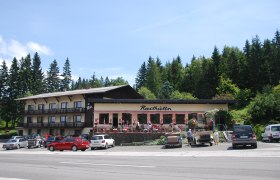 Rasthaus Brettlbar Lang, © Brettlbar Ein Rasthaus mit Terrasse und geparkten Autos davor, umgeben von Bäumen.