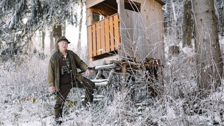 Seniorchef Karl Donhauser ist passionierter Jäger, © Niederösterreich Werbung/Mara Hohla Ein Mann in Jagdkleidung steht neben einem Hochsitz im verschneiten Wald.