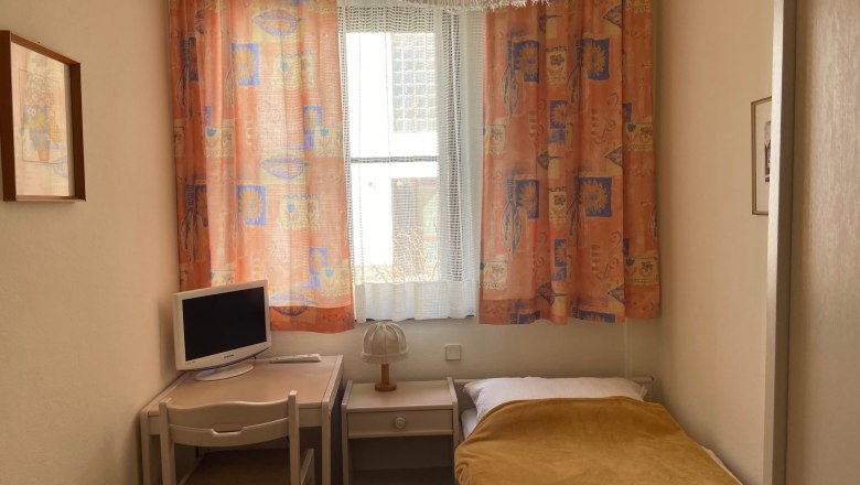 Einzelzimmer, © Wiener Alpen Ein kleines Einzelzimmer mit Bett, Schreibtisch, Stuhl und Fernseher. Bunte Vorhänge und eine Lampe sind ebenfalls zu sehen.