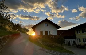 Ferienwohnung Koglhof, © Andrea Tauchner/Koglhof Ein Haus auf einem Hügel bei Sonnenuntergang mit einer kurvigen Straße im Vordergrund.