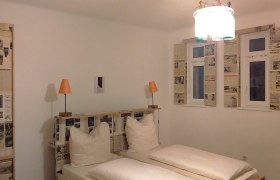 Zimmer im Hotel Binder, © Wiener Alpen Hotelzimmer mit Doppelbett, Zeitungsdekoration an Wänden und Fenstern, zwei orangefarbene Lampen.