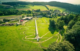 Die Wexl Arena St.Corona, © Wexl Arena St. Corona am Wechsel Luftaufnahme der Wexl Arena in St. Corona mit grünen Hügeln und verschlungenen Wegen.