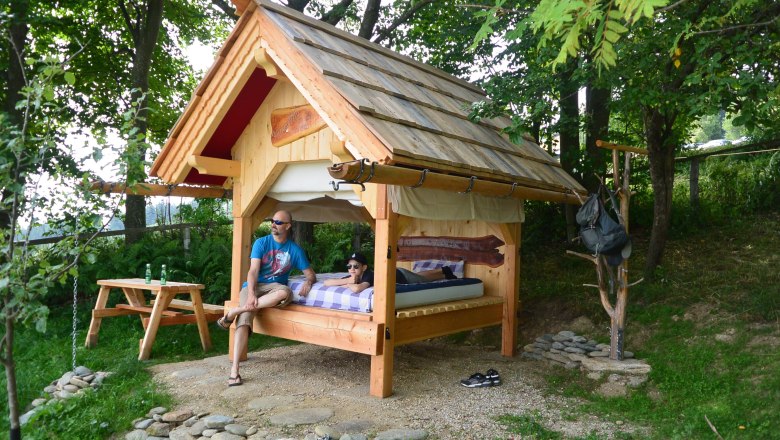 Bett im Freien in St. Corona am Wechsel, © Erika Kronaus Ein kleines Holzhaus mit einem Bett im Freien, umgeben von Bäumen. Zwei Personen sitzen und liegen auf dem Bett. Ein Picknicktisch steht daneben.