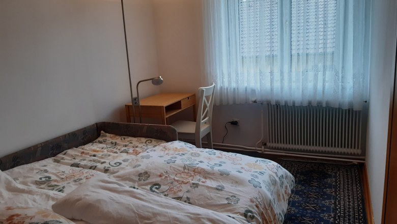 Ausziehcouch - Schlafmöglichkeit für 2 Personen, © Kainz-Waldinger Ein Zimmer mit einer ausgezogenen Couch als Bett, einem Schreibtisch mit Stuhl und einem Fenster mit Vorhängen.