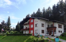 Gästehaus Apschner, © Robert Apschner Ein zweistöckiges Gästehaus mit roten Balkonen und Blumen, umgeben von Bäumen und einem blauen Himmel.