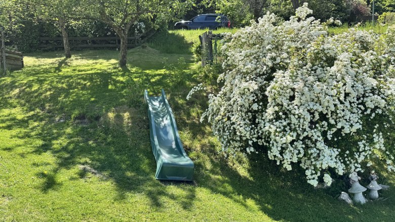 Ferienwohnung Koglhof, © Andrea Tauchner/Koglhof Grüne Rutsche auf einem Hügel im Garten, umgeben von Bäumen und blühenden Sträuchern.