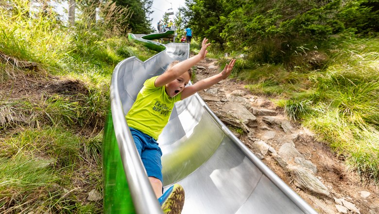 Viele Attraktionen, wie die Alm Rutsche, © Erlebnisalm Mönichkirchen, Christian Kremsl Kind rutscht auf einer Metallrutsche im Freien.