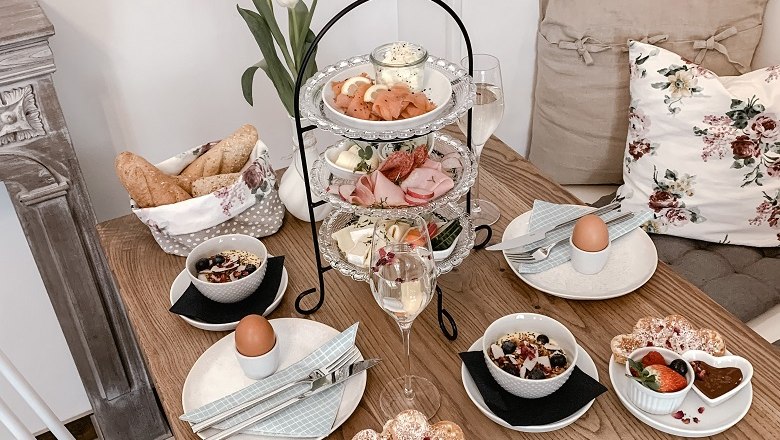 Liebevoll zubereitete Frühstücksvariationen, © Mille Fleurs Ein gedeckter Frühstückstisch mit Etagere, Brötchen, Eiern, Müsli und Obst. Dekoriert mit Tulpen und Kissen.