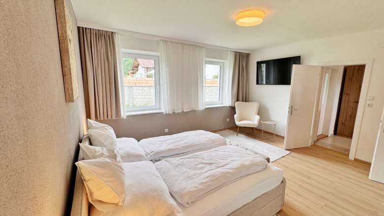 Schlafzimmer, © Christoph Menhofer Modernes Schlafzimmer mit Doppelbett, zwei Fenstern, Sessel und Fernseher an der Wand.