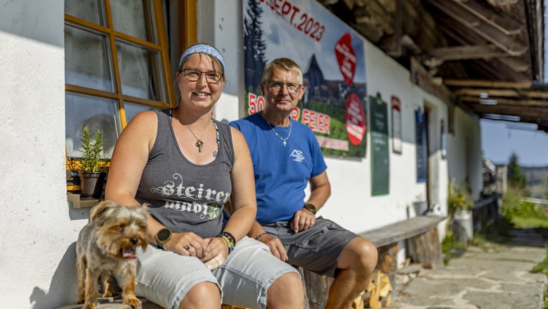 Die Wirtsleut´von der Marienseer Schwaig, © Wiener Alpen, Christian Kremsl Zwei Personen sitzen vor einem Gebäude mit einem Hund, im Hintergrund ein Plakat mit Datum 09. Sept. 2023.