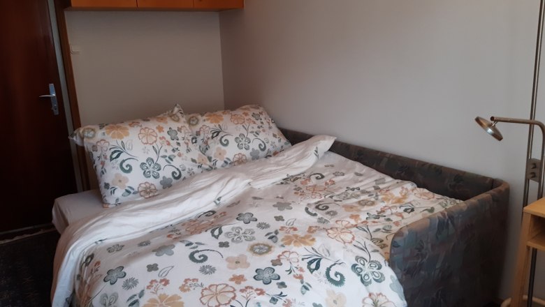 2. Zimmer - Schlafmöglichkeit für 2 Personen, © Kainz-Waldinger Ein kleines Zimmer mit einem ausziehbaren Sofa, das als Bett für zwei Personen dient, mit floraler Bettwäsche und einem Leselicht daneben.