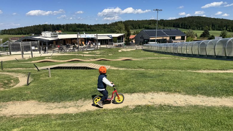 Ferienwohnung Koglhof, © Andrea Tauchner/Koglhof Ein Kind mit Helm fährt auf einem Laufrad über einen grasbewachsenen Hügel. Im Hintergrund sind Gebäude und ein überdachter Gehweg zu sehen.