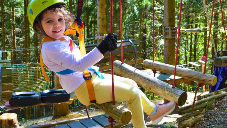 Viel Spaß am Kinderparcours, © Hamari Kletterpark Ein Kind mit Helm und Sicherheitsgurt sitzt auf einem Holzparcours im Wald.