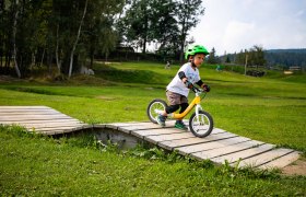 Mini Bikepark Wexl Trails, © Wexl Arena St. Corona am Wechsel Ein Kind mit grünem Helm fährt auf einem Laufrad über eine Holzrampe im Mini Bikepark Wexl Trails.