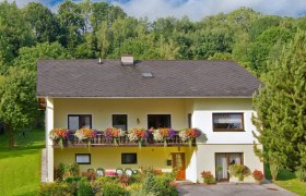 Unser Haus, © Wachabauer Zweistöckiges gelbes Haus mit Balkon und Blumen, umgeben von Bäumen und Garten.