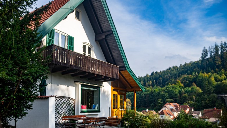 Hausansicht, © Peter Wochesländer Ein weißes Haus mit rotem Dach und grünem Balkon vor einem bewaldeten Hügel.