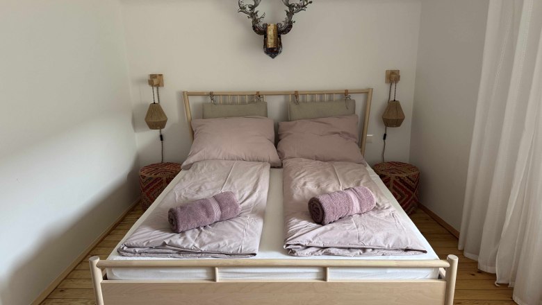 Zimmeransicht in Dottore Casa, © Michael Liska Schlafzimmer mit Doppelbett, rosa Bettwäsche und Handtüchern, Holzboden und Wanddekoration.
