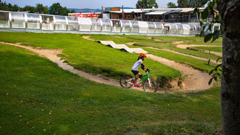 Mini Bikepark, © Wexl Arena St. Corona am Wechsel Ein Kind mit orangem Helm fährt auf einem grünen Fahrrad durch einen Mini-Bikepark mit Hügeln und Kurven.