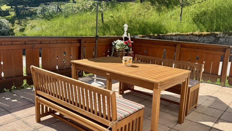 Ferienwohnung Koglhof, © Andrea Tauchner/Koglhof Holzterrasse mit Tisch, Bänken und Sonnenschirm, umgeben von grünem Garten.
