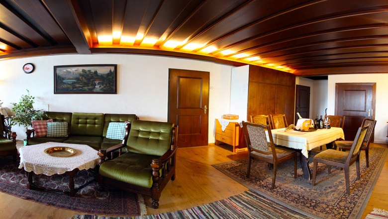 Gem&uuml;tliches Wohnzimmer mit Holzm&ouml;beln, gr&uuml;nem Sofa, Esstisch und Teppichen.