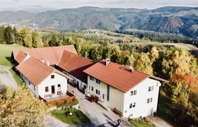 Luftaufnahme eines Bauernhofs mit roten D&auml;chern in einer h&uuml;geligen Landschaft.
