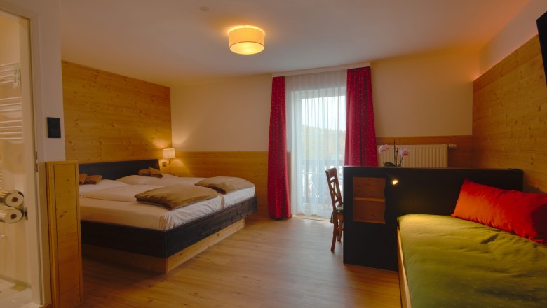 Ein gem&uuml;tliches Doppelzimmer mit Holzw&auml;nden, einem Doppelbett, einem Sofa und einem Schreibtisch. Ein Fenster mit roten Vorh&auml;ngen l&auml;sst Tageslicht herein.