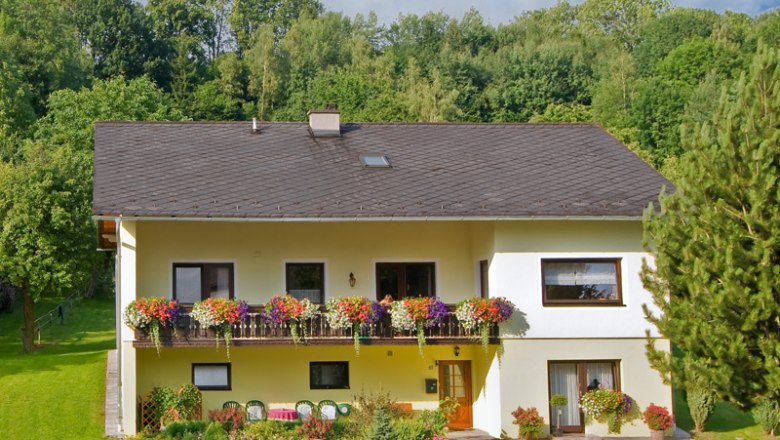 Unser Haus, © Wachabauer Zweistöckiges gelbes Haus mit Balkon und Blumen, umgeben von Bäumen und Garten.