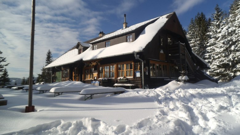 Verschneite Berghütte mit Holzbänken im Vordergrund und Tannen im Hintergrund.