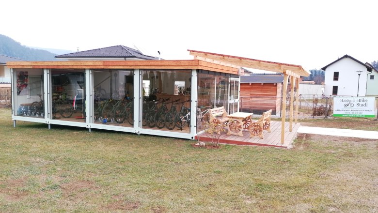 Au&szlig;enansicht eines modernen Fahrradverleihs mit Glasfront und Holzdach, umgeben von Rasen und einem Schild mit der Aufschrift 'Haiden's eBike Stadl'.