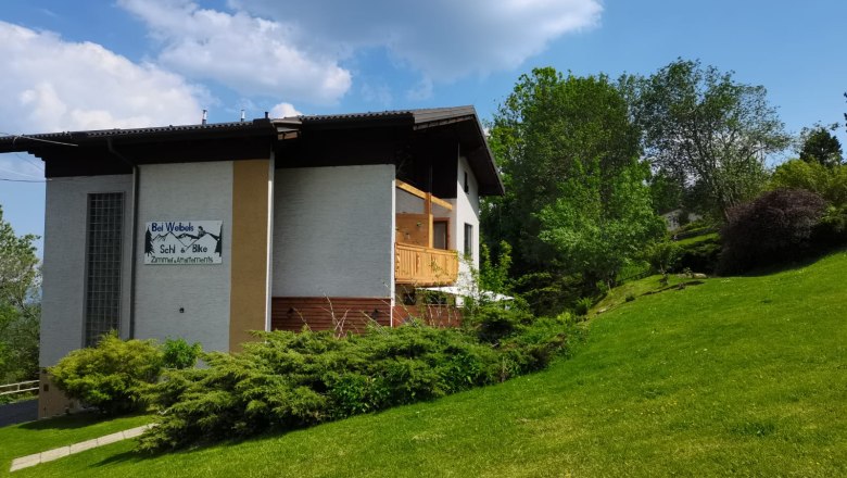 Haus mit Schild 'Bei Webels Ski & Bike' auf gr&uuml;ner Wiese, umgeben von B&auml;umen, unter blauem Himmel.