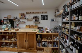 Innenansicht eines kleinen, gemütlichen Ladens mit Holzregalen, gefüllt mit Weinflaschen, Lebensmitteln und einer alten Registrierkasse.