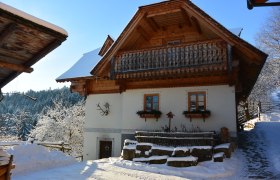 Traditionelles Haus mit Halzbalkon und weißer Fassade, umgeben von verschneiter Landschaft.