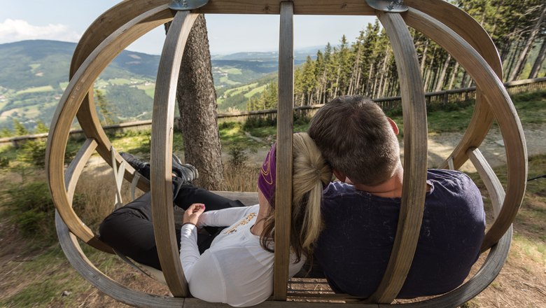 Zwei Personen sitzen in einer Holzschaukel mit Blick auf eine h&uuml;gelige Landschaft.