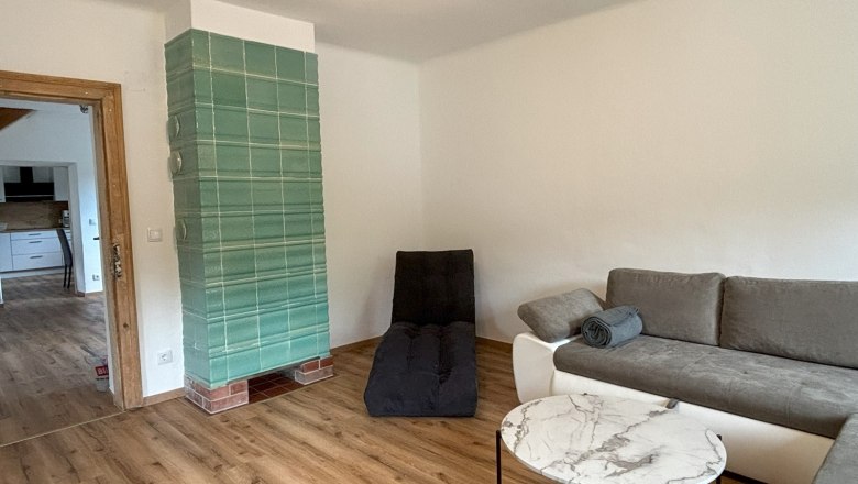 Wohnzimmer mit grauem Sofa, rundem Marmortisch und gr&uuml;nem Kachelofen.