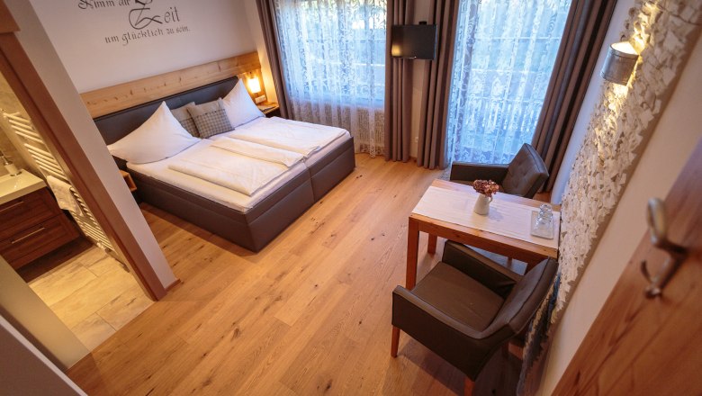 Gemütliches Doppelzimmer mit Holzboden, Doppelbett, kleinem Tisch und Stühlen, dekorativen Vorhängen und einem Fernseher an der Wand.