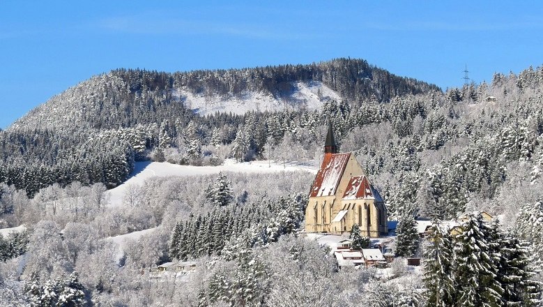 Winterlandschaft mit Kirche vor bewaldetem Hügel.