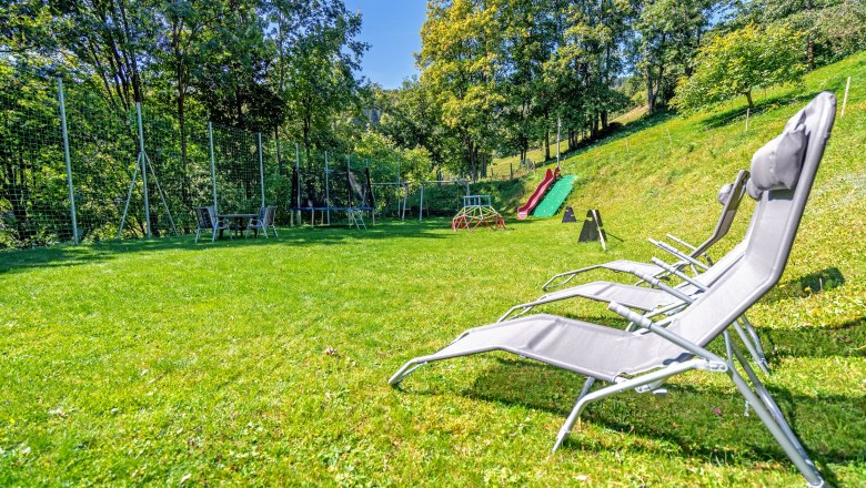 Spielplatz mit Rutsche, Trampolin und Liegestühlen auf einer Wiese, umgeben von Bäumen.