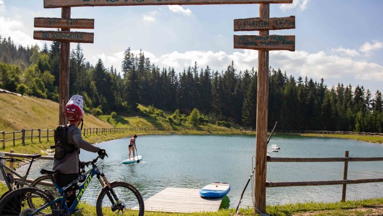 Ein Radfahrer steht vor einem Speicherteich mit SUP-Station in St. Corona am Wechsel. Eine Person paddelt auf dem Teich.