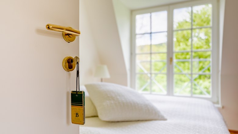 Offene Tür mit Schlüssel und Schild, dahinter ein Bett und Fenster.