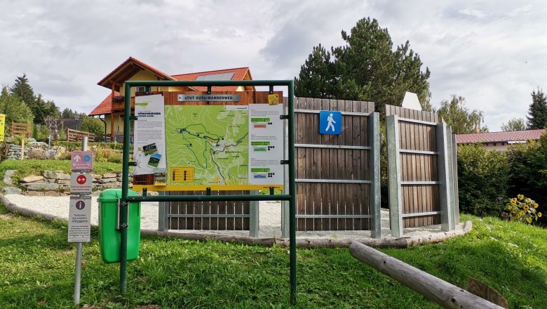 Wanderweg-Infotafel mit Karte und Hinweisschildern auf einer Wiese, umgeben von B&auml;umen und einem Geb&auml;ude im Hintergrund.