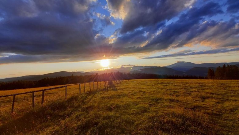 Sonnenuntergang über einer grünen Wiese mit Bergen im Hintergrund und dramatischen Wolken am Himmel.