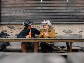 Zwei Mountainbiker sitzen auf einer Holzbank vor einer H&uuml;tte und trinken Erfrischungsgetr&auml;nke.