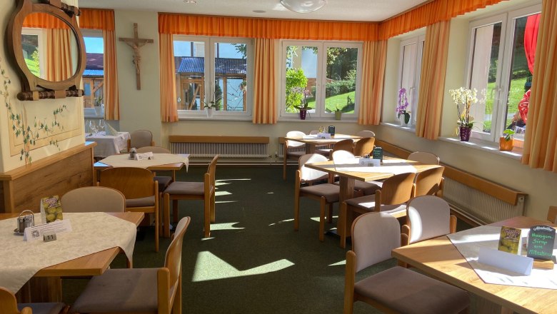 Ein heller Speisesaal mit Holztischen, Stühlen und großen Fenstern mit orangefarbenen Vorhängen.