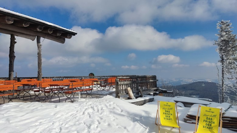 Verschneite Terrasse mit orangefarbenen Bänken und zwei gelben Liegestühlen, umgeben von schneebedeckten Bäumen und Bergen im Hintergrund.