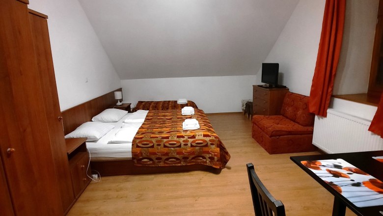 Ein einfach eingerichtetes Zimmer mit Doppelbett, Sofa, Tisch und Stuhl. Holzmöbel und orangefarbene Vorhänge dominieren den Raum.