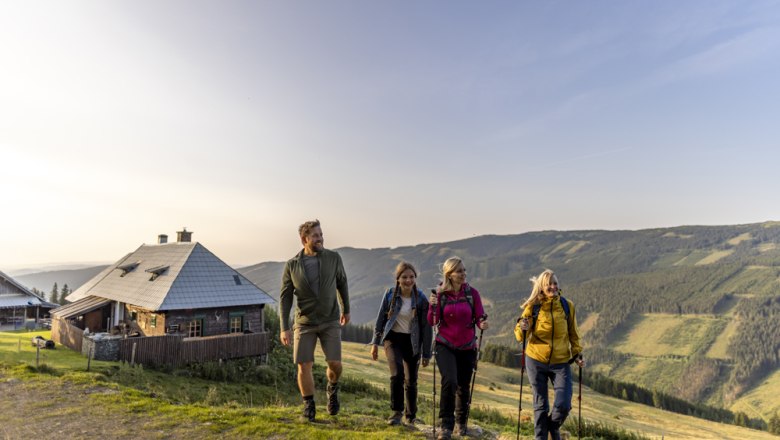 Vier Personen wandern auf einem Bergweg neben einer Hütte bei Sonnenuntergang.