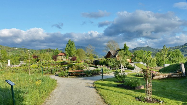 Ein gepflegter Garten mit Wegen, B&auml;umen und Blumen, umgeben von gr&uuml;nen H&uuml;geln und einem blauen Himmel mit Wolken.