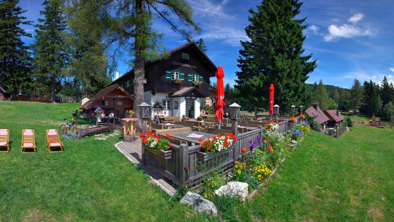 Ein Berggasthof mit Terrasse, Blumen und Liegest&uuml;hlen auf einer gr&uuml;nen Wiese, umgeben von B&auml;umen und einem klaren blauen Himmel.