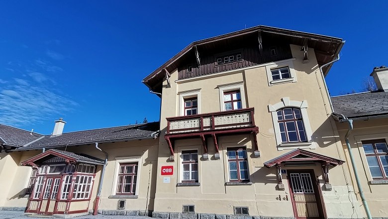 Historisches Bahnhofsgeb&auml;ude mit gelber Fassade und rotem Holzdekor unter blauem Himmel.